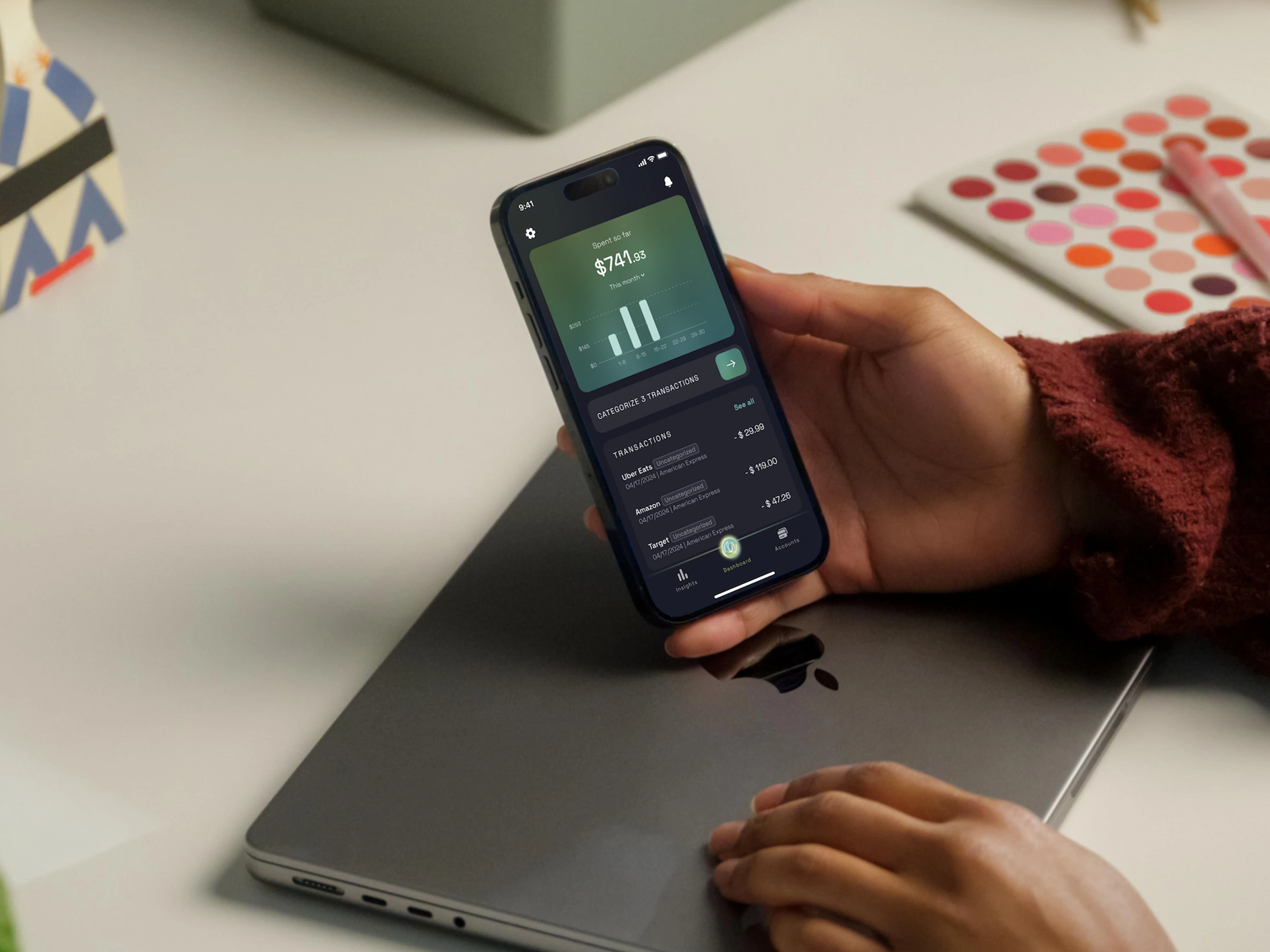 The Cashews dashboard held in one hand above a laptop, showing the amount spent so far this month, a weekly bar chart and three uncategorised transactions
